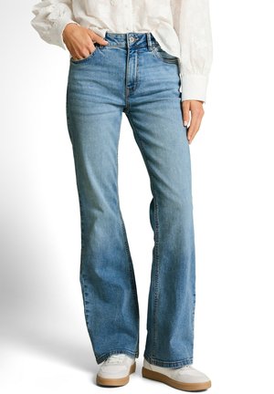 Frau trägt hellblaue, hoch taillierte Schlagjeans, weiße Turnschuhe und eine weiße Langarmbluse, mit einer Hand in der Tasche.