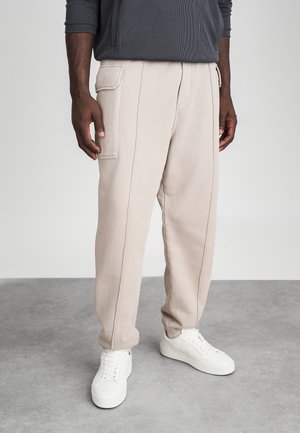 Béžové tepláky so voľným strihom, vertikálnymi švami a bočnými vreckami, kombinované s bielymi teniskami. Hladká textúra s neformálnym dizajnom.