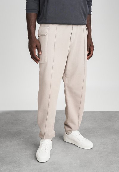 Sudaderas beige de corte holgado, con costuras verticales y bolsillos laterales, combinadas con zapatillas blancas. Textura suave con un diseño casual.