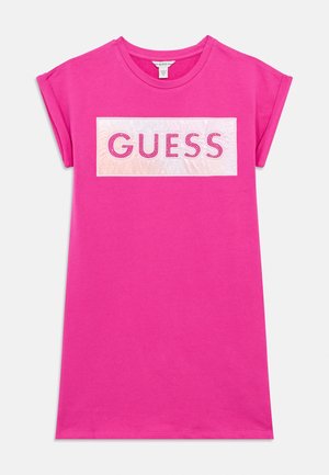 Felle roze jurk met korte mouwen en een wit rechthoekig vlak op de borst met het woord "GUESS" in dikke roze letters.
