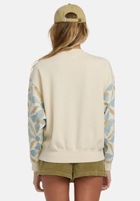 Sudadera de crema claro con patrones de hojas azules y doradas en las mangas, puños acanalados y un ajuste relajado. Pantalones cortos de pana cortos a continuación.