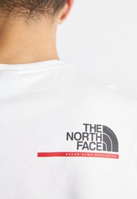 Bílý bavlněný tričko s černým a šedým logem "THE NORTH FACE" a červeným nápisem "NEVER STOP EXPLORING" na zadním límci.