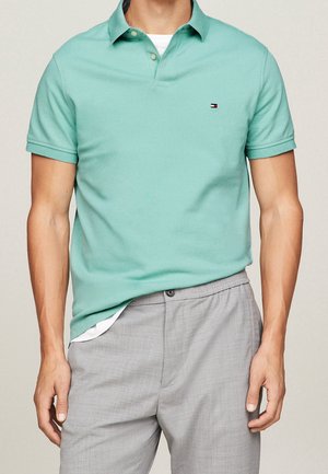 Uomo che indossa una polo a maniche corte verde chiaro sopra una maglietta bianca, con pantaloni grigio chiaro, in piedi contro uno sfondo semplice.