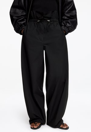 Pantalon noir à jambes larges avec taille élastiquée et cordon, porté avec une veste en cuir noire et des tongs noires pieds nus.