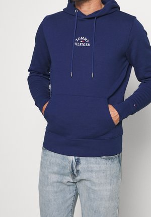 Mann trägt eine dunkelblaue Tommy Hilfiger Kapuzenjacke mit Kängurutasche und hellblaue Jeans, Hände in den Taschen der Kapuzenjacke, vor einem schlichten Hintergrund.