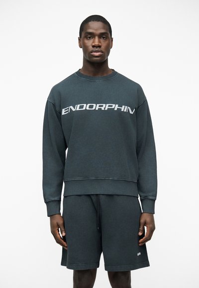 Homme portant un sweat-shirt gris foncé avec le texte "ENDORPHIN" et un short assorti, debout devant un fond blanc uni.