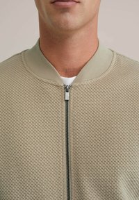 Chaqueta bomber texturizada en color beige con cierre de cremallera, cuello acanalado y un distintivo patrón de panal. Tejido suave con un brillo sutil.