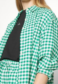 Green and white gingham button-up shirt with a collar, featuring short sleeves and a black undershirt. Includes a small silver heart-shaped pin.