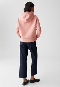 Persona in piedi di spalle con indosso un hoodie rosa chiaro, jeans cropped blu scuro e scarpe slip-on nere su uno sfondo grigio uniforme.