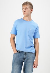 Világoskék pamut póló kerek nyakkal és rövid ujjal, bal oldalon zsebbel és laza szabással. Világoskék farmernadrággal viselve.