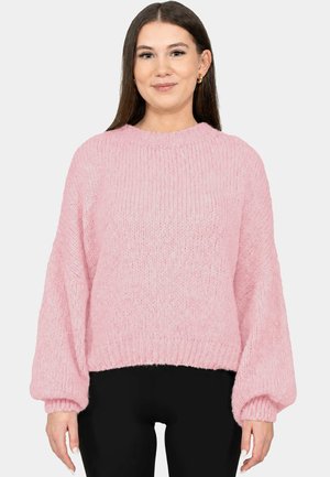Woman with long brown hair wearing gold hoop earrings and a loose pink knitted sweater with balloon sleeves, paired with black pants.