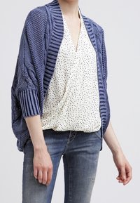 Cardigan de punto en azul oscuro con texturas variadas, de frente abierto y hombros caídos, superpuesto sobre una blusa blanca con lunares negros.