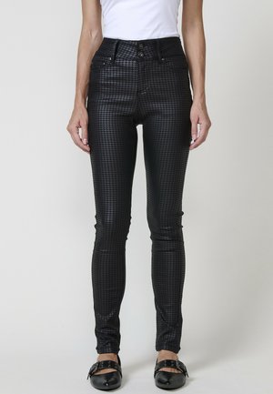 Pantalon en cuir noir slim avec motif pied-de-poule embossé, associé à des chaussures noires à boucle et un top blanc sans manches.