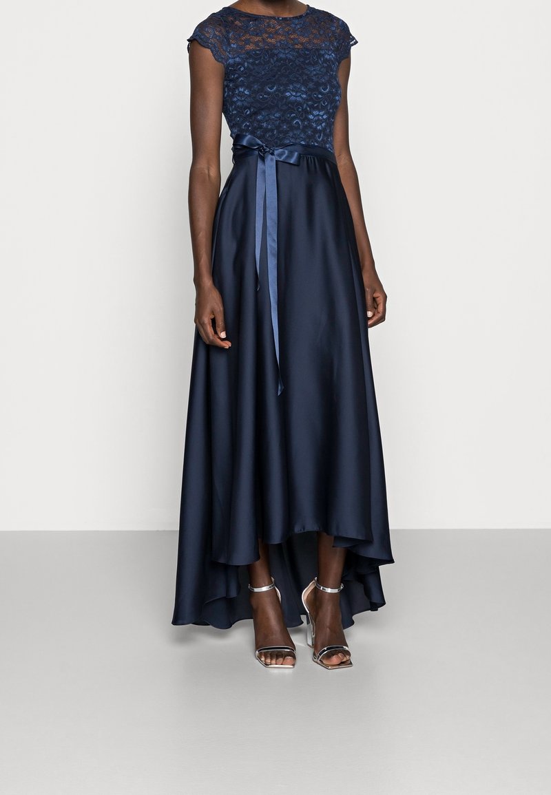 Femme portant une robe de soirée bleu marine en dentelle et satin à ourlet asymétrique avec un ruban à la taille, associée à des talons hauts argentés avec bride à la cheville.