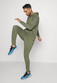 Olivenfarbener Hoodie und passende Jogginghose aus weichem Stoff. Die Jogginghose hat eine Cargo-Tasche. Blaue Sportschuhe mit schwarzen Akzenten.