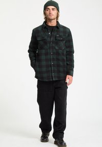 Green and black plaid flannel shirt with two chest pockets, button closure, worn over a black shirt, paired with black cargo pants and black boots.