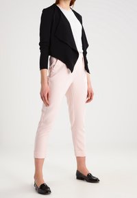 Veste noire drapée sur un haut blanc, associée à un pantalon tapered rose clair et des ballerines noires brillantes. Le tissu semble lisse ; le design est moderne.