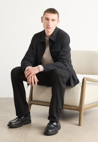 Black textured jacket over a beige top, paired with black trousers and black shoes. The model sits on a light-colored chair against a plain background.
