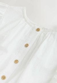 Balta kokvilnas blūze ar apaļu kakla izgriezumu, tai ir pogu plāksne ar koka pogām un teksturētām ziedu rakstiem.