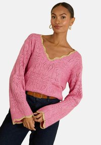 Roze gebreide trui met een V-hals, een ingewikkeld kantpatroon en een gekartelde gele boord bij de kraag en mouwen. Lange klokvormige mouwen.