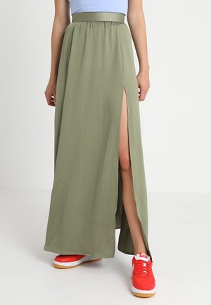 Kvinde iført olivengrønt maxi-nederdel med benhøjt slids og skrigrøde sneakers, stående mod en hvid baggrund.