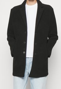 Manteau en mélange de laine noir avec revers crantés, fermeture à cinq boutons et poches latérales, superposé à un t-shirt blanc et un jean bleu clair.
