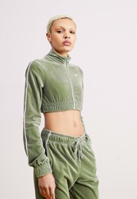 Chándal de terciopelo verde que incluye una chaqueta corta con cremallera y cuello alto, junto con pantalones de cintura elástica a juego, acentuados con rayas blancas.