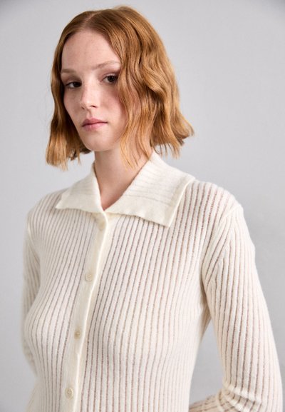 Cardigan cu textură ribbată, de culoare crem, cu guler ascuțit, având închidere cu nasturi și o textură moale, așezat pe un fundal neutru.