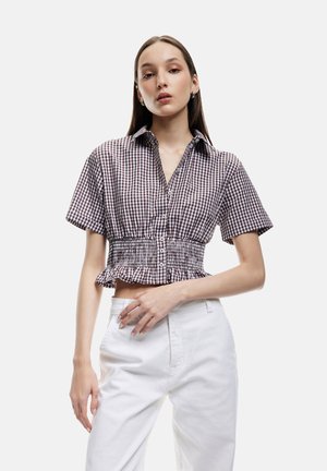 Camicia a quadretti marrone e bianca, grazie alla sua forma cropped e alle maniche corte, con vita arricciata e chiusura a bottoni. Abbinata a pantaloni bianchi a vita alta.