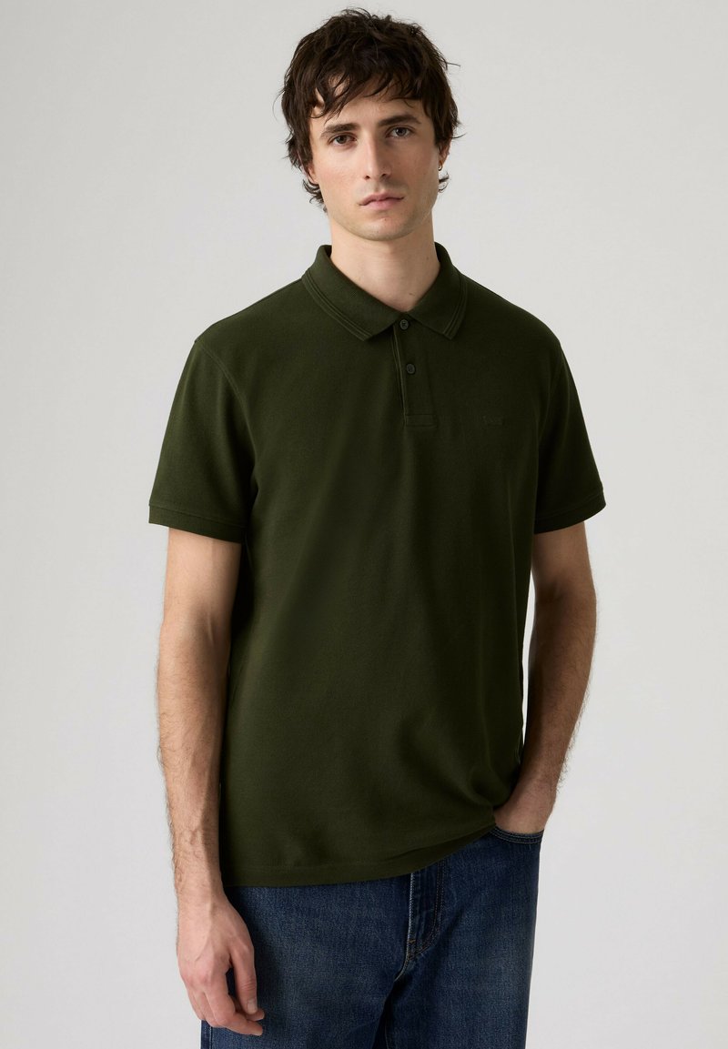Jeune homme aux cheveux courts et foncés, portant un polo vert olive et un jean bleu, debout avec une main dans la poche devant un fond uni.