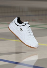 Weißer Sportschuh mit strukturiertem Obermaterial, schwarzen Akzenten und einer Sohle aus Gummilaufsohle; verfügt über Schnürsenkel und ein Logo an der Seite.