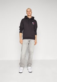 Sudadera con capucha negra, con bolsillo frontal y detalle de logo, combinada con jeans gris claro y zapatillas blancas. Material de algodón suave, corte relajado.