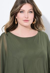 Top leggero e fluido di colore verde oliva, con scollatura ampia e maniche trasparenti. Sono visibili grandi orecchini a cerchio tondeggianti in tono dorato.
