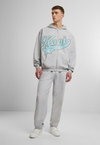 Pelēks zipsvārku džemperis ar zilu "Kani" emblēmu un zvaigžņu dizainu, apvienots ar atbilstošām sporta biksēm un baltām kedām. Kvalitatīvs kokvilnas auduma maisījums.
