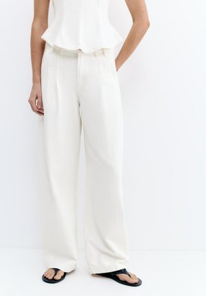 Kobieta w białych spodniach z wysokim stanem i szerokimi nogawkami oraz czarnych klapkach stojąca na jednolitym białym tle.