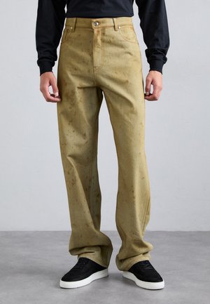 Pantaloni larghi color sabbia con una finitura testurizzata, caratterizzati da macchie visibili, una chiusura a bottoni e cuciture arancioni; indossati con sneakers nere.