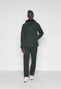 Donkergroen waterdicht jack met capuchon, gecombineerd met bijpassende cargobroek. Draagbaar met een groene muts en witte zonnebril, afgewerkt met lichte sneakers.