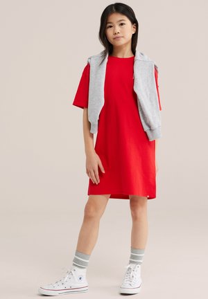 Jauna meitene stāv, valkājot sarkanu kleitu ar īsām piedurknēm, baltas augstcilvēku kedas, pelēkas svītrainas zeķes un pelēku džemperi, kas pārmetts pāri pleciem.