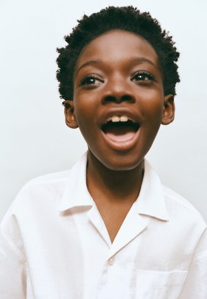 Mladý černoch s krátkymi kučeravými vlasmi, oblečený v bielej košeli, s otvorenými ústami, akoby spieval alebo hovoril, na jednolivom svetlom pozadí.