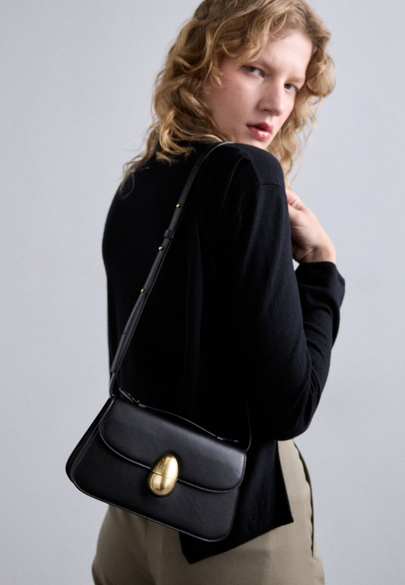 Black leather handbag with a curved shape, featuring a prominent gold oval clasp, adjustable strap, and minimalistic design.