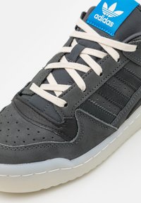 Grå sneakers med en blandning av slät och texturerad läder, perforerade detaljer, gräddfärgade snörer och en blå Adidas-logotyp på tungan.