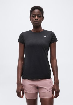 Femeie purtând un tricou negru mulat cu mâneci scurte și pantaloni scurți sport roz, stând în picioare pe un fundal simplu și deschis la culoare.