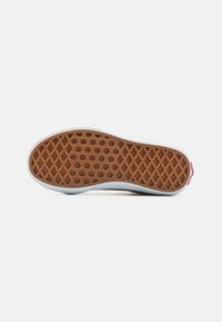 Semelle d'une sneaker avec une base en caoutchouc marron, dotée d'une texture à motif en losange, bordée de blanc et d'un accent rouge au talon.