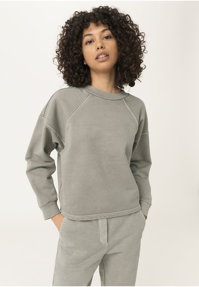 Sudadera gris clara hecha de una tela suave, con cuello redondo, mangas raglán y puños acanalados. Combinada con pantalones de chándal a juego.