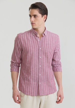 Jeune homme portant une chemise rose à manches longues avec des rayures blanches verticales et un pantalon beige, regardant vers sa gauche sur un fond uni.