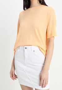 Vrouw in een perzikkleurig T-shirt met korte mouwen, gestopt in een witte spijkerrok met voorzakken en knoopsluiting, staand met één arm ontspannen.