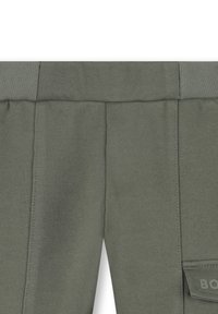 Groene, gestructureerde stofbroek met elastische tailleband en gedeeltelijk zichtbare zijzakflap met reliëftekst.