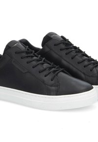 Baskets en cuir noir avec semelles en caoutchouc blanches, présentant des bouts ronds, des lacets plats noirs et un design minimaliste. Détails du logo sur les côtés.