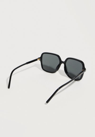 Γυαλιά ηλίου με μαύρο τετράγωνο πλαίσιο, σκουρόχρωνους φακούς, λεπτά πλαϊνά και διακριτικές χρυσές λεπτομέρειες στα μεντεσέδες, τοποθετημένα σε λευκή επιφάνεια.