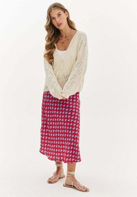 Pletený krémový cardigan s vzormi srdca, navrstvený na červenej sukni s modrými abstraktnými tvarmi, doplnený hnedými sandálmi na remienky.